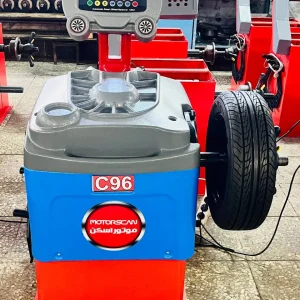 A wheel balancing machine labeled "C96" with the brand "MOTORSCAN" written in both English and Arabic is positioned in a workshop. The machine features a digital display with multiple buttons and indicators, allowing for precise wheel balancing. A tire is mounted on the right side, ready for adjustments. The background contains other similar equipment, reinforcing the professional automotive setting.