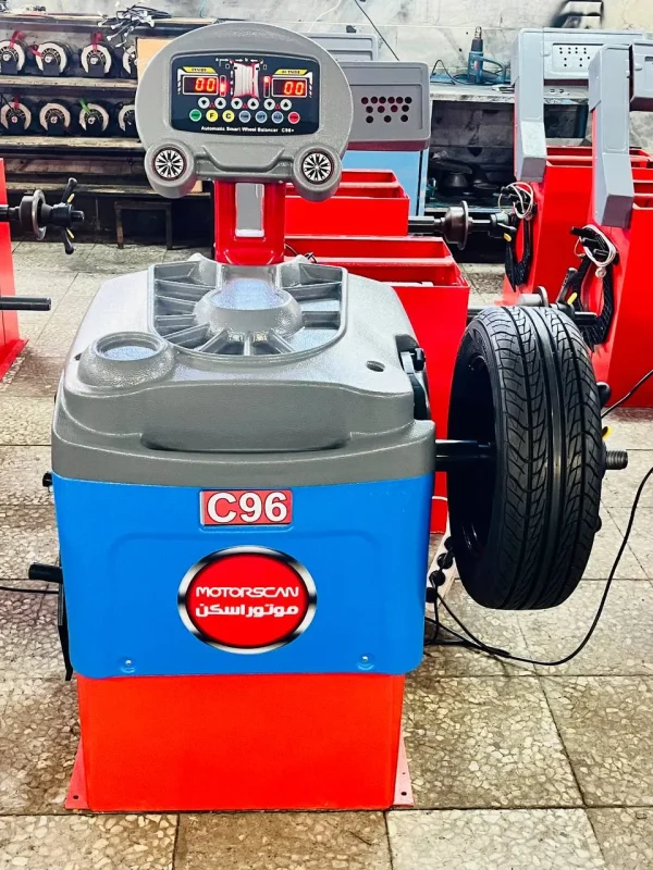 A wheel balancing machine labeled "C96" with the brand "MOTORSCAN" written in both English and Arabic is positioned in a workshop. The machine features a digital display with multiple buttons and indicators, allowing for precise wheel balancing. A tire is mounted on the right side, ready for adjustments. The background contains other similar equipment, reinforcing the professional automotive setting.