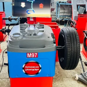 A tire balancing machine labeled "M97" with the brand name "MOTORSCAN" written in both English and Arabic stands in a workshop. A tire is mounted on the machine, and its screen displays a graphical representation of the tire's balance status. The diagram on the screen includes arrows indicating necessary adjustments for proper balance. In the background, additional similar machines and equipment reinforce the professional automotive setting.