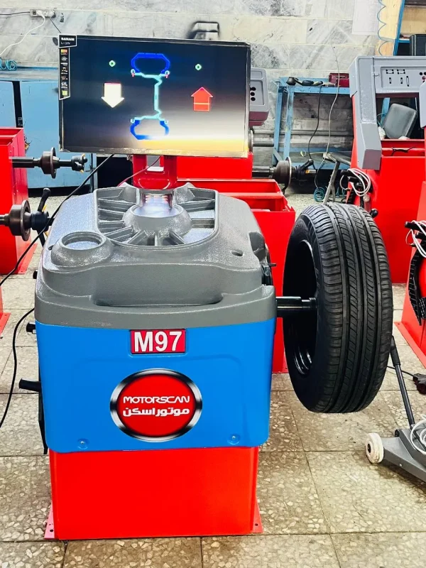 A tire balancing machine labeled "M97" with the brand name "MOTORSCAN" written in both English and Arabic stands in a workshop. A tire is mounted on the machine, and its screen displays a graphical representation of the tire's balance status. The diagram on the screen includes arrows indicating necessary adjustments for proper balance. In the background, additional similar machines and equipment reinforce the professional automotive setting.
