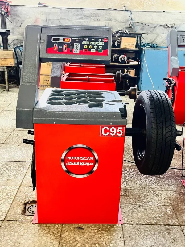 A red and gray machine labeled "C95" with the brand name "MOTORSCAN" written in both English and Arabic stands in a workshop. The machine appears to be a smart wheel balancer, used for ensuring vehicle wheels are properly balanced. Its control panel features various buttons and a digital display showing "0.0." A wheel is mounted on the right side, ready for balancing. The background includes other automotive equipment and tools, reinforcing the professional workshop setting.