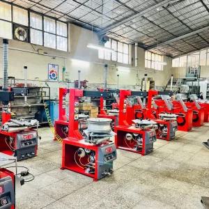 The image shows an industrial workshop with multiple red MT110 machines lined up in rows. These machines appear to be tire changers or similar equipment used for automotive purposes. The workshop has a high ceiling with metal roofing and large windows that allow natural light to enter. There are various tools and equipment scattered around, including a fan on the wall, boxes, and other machinery. The floor is tiled, and the overall environment is clean and organized.