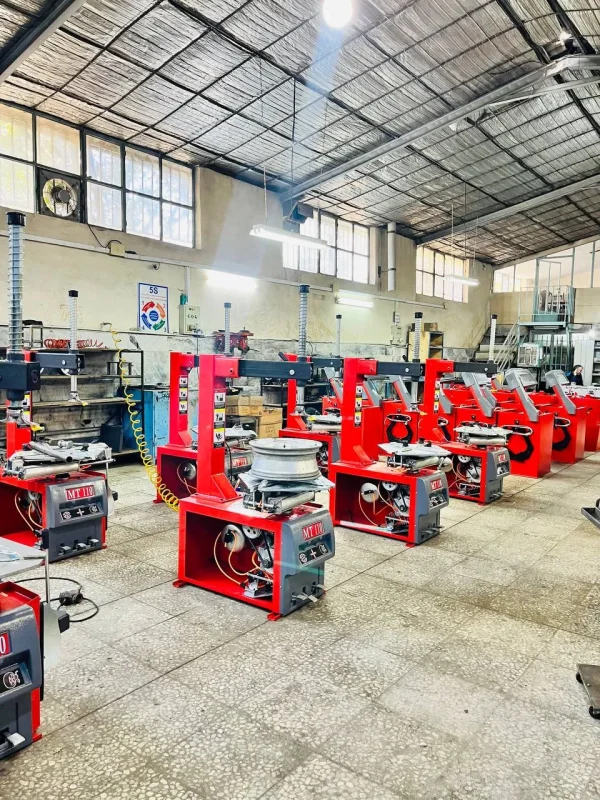 The image shows an industrial workshop with multiple red MT110 machines lined up in rows. These machines appear to be tire changers or similar equipment used for automotive purposes. The workshop has a high ceiling with metal roofing and large windows that allow natural light to enter. There are various tools and equipment scattered around, including a fan on the wall, boxes, and other machinery. The floor is tiled, and the overall environment is clean and organized.