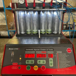 A digital fuel injector cleaner and tester machine labeled "S700+ Automatic Injector Cleaner & Tester" is set up for operation. The machine features a digital display with various settings, buttons, and a menu listing multiple testing functions. Six transparent cylinders filled with a green liquid are connected to red clamps and blue wires, allowing for measurement and analysis. The control panel includes a pressure gauge and connectors labeled for different testing functions. The interface presents data such as pulse width, working time, and injection values, making it a sophisticated tool for injector diagnostics.