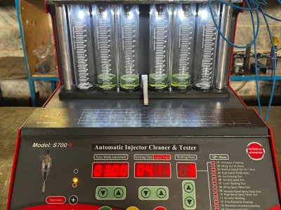 A digital fuel injector cleaner and tester machine labeled "S700+ Automatic Injector Cleaner & Tester" is set up for operation. The machine features a digital display with various settings, buttons, and a menu listing multiple testing functions. Six transparent cylinders filled with a green liquid are connected to red clamps and blue wires, allowing for measurement and analysis. The control panel includes a pressure gauge and connectors labeled for different testing functions. The interface presents data such as pulse width, working time, and injection values, making it a sophisticated tool for injector diagnostics.