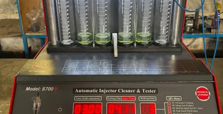 A digital fuel injector cleaner and tester machine labeled "S700+ Automatic Injector Cleaner & Tester" is set up for operation. The machine features a digital display with various settings, buttons, and a menu listing multiple testing functions. Six transparent cylinders filled with a green liquid are connected to red clamps and blue wires, allowing for measurement and analysis. The control panel includes a pressure gauge and connectors labeled for different testing functions. The interface presents data such as pulse width, working time, and injection values, making it a sophisticated tool for injector diagnostics.