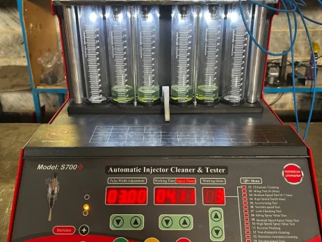 A digital fuel injector cleaner and tester machine labeled "S700+ Automatic Injector Cleaner & Tester" is set up for operation. The machine features a digital display with various settings, buttons, and a menu listing multiple testing functions. Six transparent cylinders filled with a green liquid are connected to red clamps and blue wires, allowing for measurement and analysis. The control panel includes a pressure gauge and connectors labeled for different testing functions. The interface presents data such as pulse width, working time, and injection values, making it a sophisticated tool for injector diagnostics.