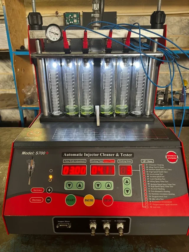 A digital fuel injector cleaner and tester machine labeled "S700+ Automatic Injector Cleaner & Tester" is set up for operation. The machine features a digital display with various settings, buttons, and a menu listing multiple testing functions. Six transparent cylinders filled with a green liquid are connected to red clamps and blue wires, allowing for measurement and analysis. The control panel includes a pressure gauge and connectors labeled for different testing functions. The interface presents data such as pulse width, working time, and injection values, making it a sophisticated tool for injector diagnostics.