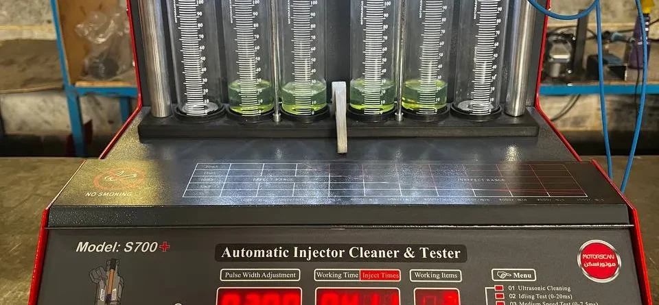 A digital fuel injector cleaner and tester machine labeled "S700+ Automatic Injector Cleaner & Tester" is set up for operation. The machine features a digital display with various settings, buttons, and a menu listing multiple testing functions. Six transparent cylinders filled with a green liquid are connected to red clamps and blue wires, allowing for measurement and analysis. The control panel includes a pressure gauge and connectors labeled for different testing functions. The interface presents data such as pulse width, working time, and injection values, making it a sophisticated tool for injector diagnostics.