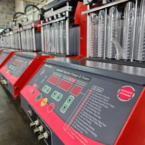 A row of machines labeled "Automatic Injector Cleaner & Tester" is arranged in a workshop setting. These machines are used for cleaning and testing fuel injectors, featuring control panels with buttons, indicators, and a detailed list of functions on the right side. Transparent tubes and gauges are integrated into the equipment, allowing users to monitor the cleaning and testing processes. The list of functions includes various tests, such as leakage detection, spray analysis, and dynamic flow assessment, emphasizing the precision involved in fuel injector maintenance.