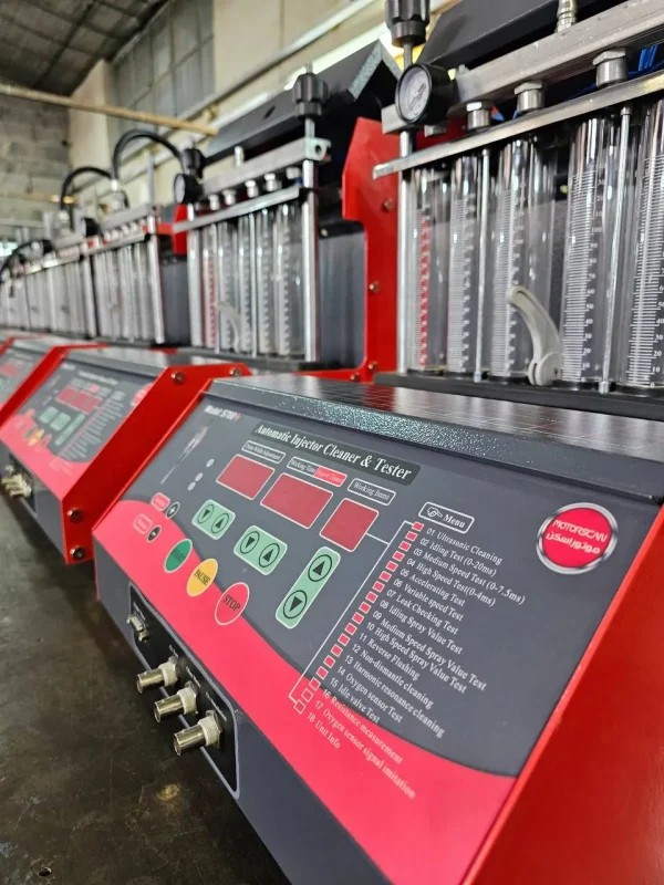 A row of machines labeled "Automatic Injector Cleaner & Tester" is arranged in a workshop setting. These machines are used for cleaning and testing fuel injectors, featuring control panels with buttons, indicators, and a detailed list of functions on the right side. Transparent tubes and gauges are integrated into the equipment, allowing users to monitor the cleaning and testing processes. The list of functions includes various tests, such as leakage detection, spray analysis, and dynamic flow assessment, emphasizing the precision involved in fuel injector maintenance.