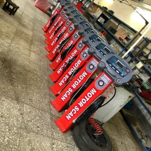A row of red diagnostic machines labeled "MOTOR SCAN" is lined up in a workshop. Each machine features a digital screen, buttons, and dials, used for motor testing and diagnostics. A tire with an orange air hose is connected to one of the machines in the foreground. Shelves filled with tools and equipment can be seen in the background, reinforcing the automotive repair setting.
