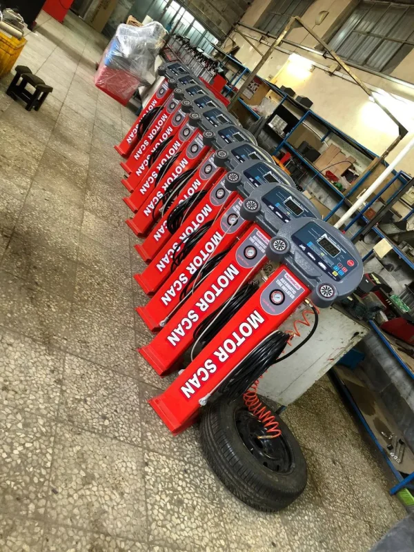 A row of red diagnostic machines labeled "MOTOR SCAN" is lined up in a workshop. Each machine features a digital screen, buttons, and dials, used for motor testing and diagnostics. A tire with an orange air hose is connected to one of the machines in the foreground. Shelves filled with tools and equipment can be seen in the background, reinforcing the automotive repair setting.