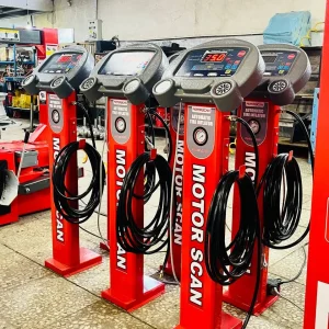 Four red automatic tire inflators from the brand MOTORSCAN are lined up in a workshop setting. Each inflator features a digital display and control panel, with coiled black hoses hanging from the front. The displays show different readings, including “35.0” and “0.0.” Various tools and equipment are visible in the background, reinforcing the professional automotive setting. The inflators are labeled “MOTORSCAN AUTOMATIC TIRE INFLATOR