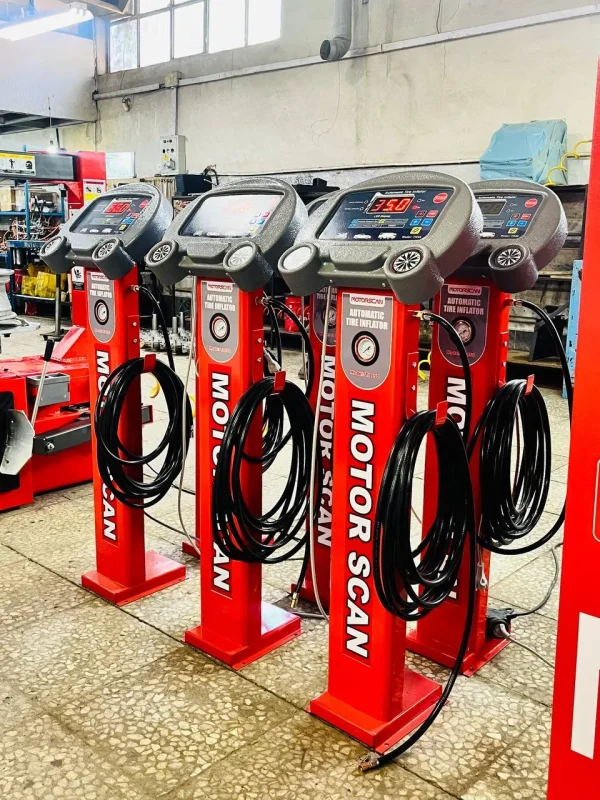 Four red automatic tire inflators from the brand MOTORSCAN are lined up in a workshop setting. Each inflator features a digital display and control panel, with coiled black hoses hanging from the front. The displays show different readings, including “35.0” and “0.0.” Various tools and equipment are visible in the background, reinforcing the professional automotive setting. The inflators are labeled “MOTORSCAN AUTOMATIC TIRE INFLATOR