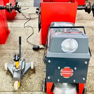 A workshop with various pieces of machinery used for automotive or mechanical work. The central focus is a machine labeled "MOTORSCAN" with Arabic text underneath, likely a brake disc lathe for resurfacing discs. To the left is a wheeled tool, possibly a hydraulic jack, and in the background, red machines with rotating components are visible. The floor is tiled, with cables and wires connecting the equipment.