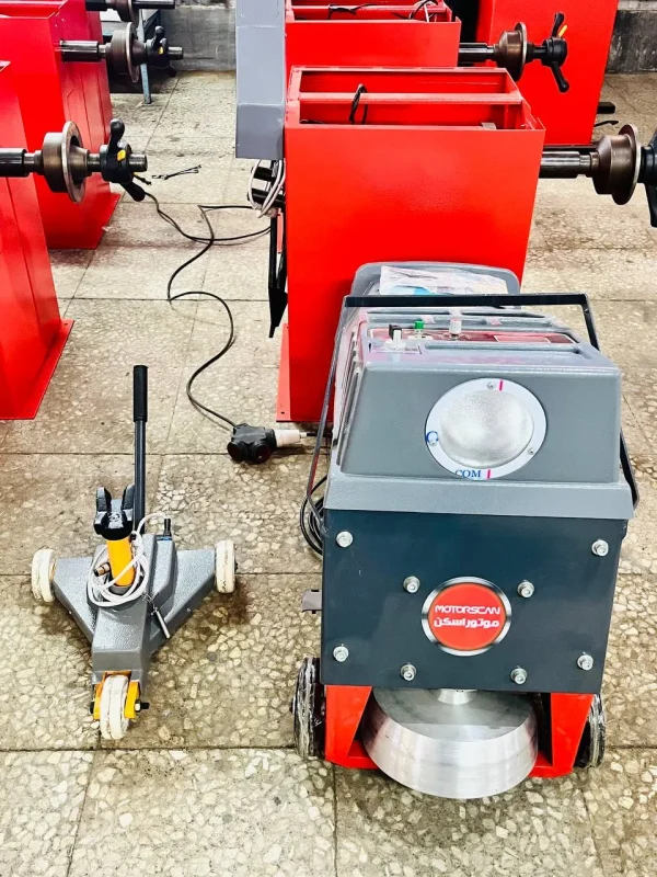 A workshop with various pieces of machinery used for automotive or mechanical work. The central focus is a machine labeled "MOTORSCAN" with Arabic text underneath, likely a brake disc lathe for resurfacing discs. To the left is a wheeled tool, possibly a hydraulic jack, and in the background, red machines with rotating components are visible. The floor is tiled, with cables and wires connecting the equipment.
