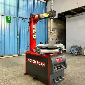 A tire changing machine labeled "MOTOR SCAN MT 190" stands in an automotive workshop. The machine has a red and black color scheme, featuring a vertical arm and a horizontal arm equipped with a tire mounting/demounting head. Foot pedals at the base control its operation. In the background, a blue metal door with an "Emergency Exit" sign is visible, along with a partially visible upper level with windows. Some equipment and parts are stacked against the wal