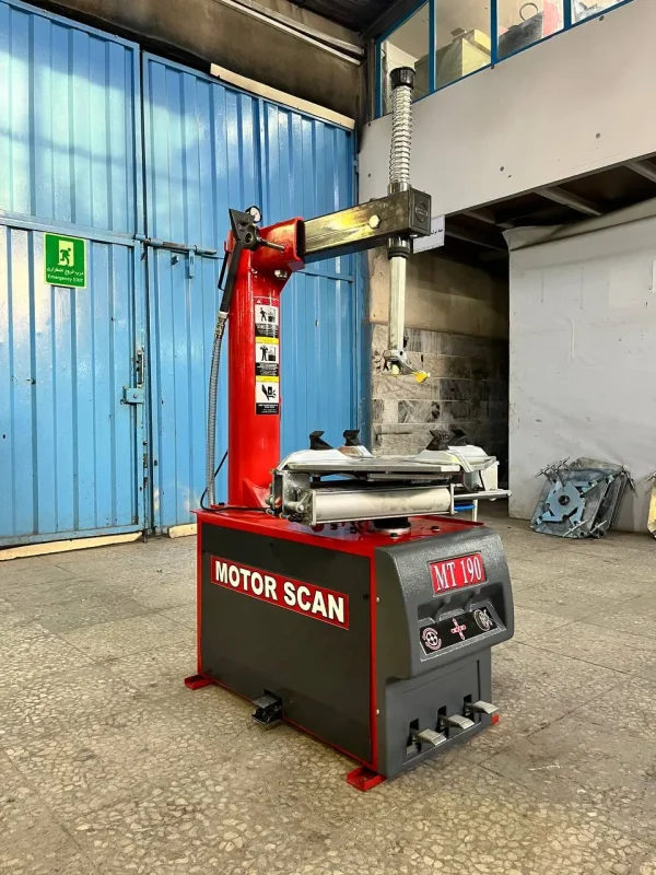 A tire changing machine labeled "MOTOR SCAN MT 190" stands in an automotive workshop. The machine has a red and black color scheme, featuring a vertical arm and a horizontal arm equipped with a tire mounting/demounting head. Foot pedals at the base control its operation. In the background, a blue metal door with an "Emergency Exit" sign is visible, along with a partially visible upper level with windows. Some equipment and parts are stacked against the wal