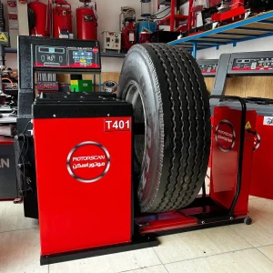 "A large tire is mounted on a red and black MOTORSCAN T401 tire balancing machine in an automotive workshop. The machine features a digital display and control buttons, with branding in both English and Arabic. Around the workspace, shelves hold various tools and equipment, including fire extinguishers and another digital machine. The setup suggests a professional environment focused on vehicle maintenance.