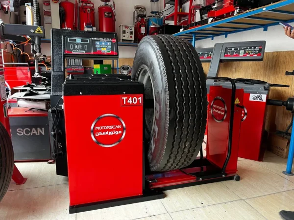 "A large tire is mounted on a red and black MOTORSCAN T401 tire balancing machine in an automotive workshop. The machine features a digital display and control buttons, with branding in both English and Arabic. Around the workspace, shelves hold various tools and equipment, including fire extinguishers and another digital machine. The setup suggests a professional environment focused on vehicle maintenance.
