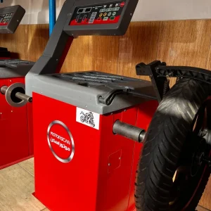 A red MOTOSCAN tire balancing machine, used for motorcycle tires, stands against a wooden wall with blue metal shelving in the background. The machine features a digital control panel with buttons and indicators, including start and stop functions. A motorcycle tire is mounted on the machine, and the label displays the brand name 'MOTOSCAN' alongside Arabic text. A small sticker with a motorcycle graphic is also visible on the machine.