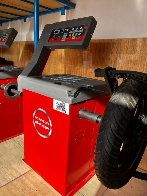 A red MOTOSCAN tire balancing machine, used for motorcycle tires, stands against a wooden wall with blue metal shelving in the background. The machine features a digital control panel with buttons and indicators, including start and stop functions. A motorcycle tire is mounted on the machine, and the label displays the brand name 'MOTOSCAN' alongside Arabic text. A small sticker with a motorcycle graphic is also visible on the machine.