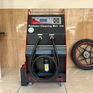 The image shows a radiator cleaning machine labeled "RC-730 Radiator Cleaning Machine." The machine has a digital display and control buttons on the top panel. Below the panel, there are two labeled ports, "INPUT" and "OUTPUT," with hoses attached to them. A pressure gauge is also visible on the left side of the machine. The machine is mounted on wheels for mobility. In the background, there is a tire with a red rim leaning against the wall. The machine is placed on a tiled floor with a wooden panel wall behind it. The text "Engine Cleaning" is partially visible at the bottom of the machine.