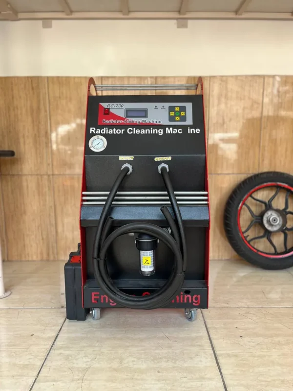The image shows a radiator cleaning machine labeled "RC-730 Radiator Cleaning Machine." The machine has a digital display and control buttons on the top panel. Below the panel, there are two labeled ports, "INPUT" and "OUTPUT," with hoses attached to them. A pressure gauge is also visible on the left side of the machine. The machine is mounted on wheels for mobility. In the background, there is a tire with a red rim leaning against the wall. The machine is placed on a tiled floor with a wooden panel wall behind it. The text "Engine Cleaning" is partially visible at the bottom of the machine.
