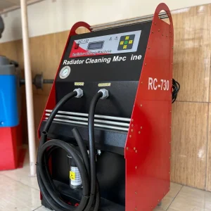 The image shows a red and black machine labeled "Radiator Cleaning Machine" with the model number "RC-730." The machine has a digital display, control buttons, and two hoses labeled "Input" and "Output." The hoses are coiled and attached to the front of the machine. The machine is placed on a tiled floor against a wooden wall.