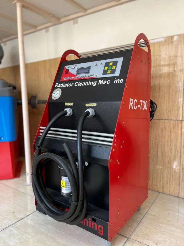 The image shows a red and black machine labeled "Radiator Cleaning Machine" with the model number "RC-730." The machine has a digital display, control buttons, and two hoses labeled "Input" and "Output." The hoses are coiled and attached to the front of the machine. The machine is placed on a tiled floor against a wooden wall.