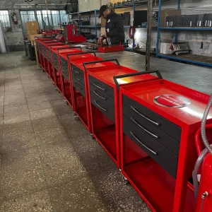 A well-organized workshop featuring several red tool carts arranged neatly in a row. Each cart has three drawers and a handle for easy mobility. A person is actively working at a bench in the background, surrounded by various tools and equipment. Shelves with organized items, a clock on the wall, and a fire extinguisher add to the professional workspace. The floor is tiled, and the environment appears clean, structured, and functional.