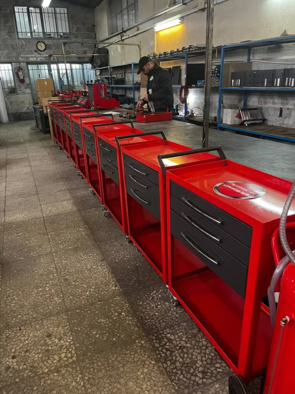 A well-organized workshop featuring several red tool carts arranged neatly in a row. Each cart has three drawers and a handle for easy mobility. A person is actively working at a bench in the background, surrounded by various tools and equipment. Shelves with organized items, a clock on the wall, and a fire extinguisher add to the professional workspace. The floor is tiled, and the environment appears clean, structured, and functional.