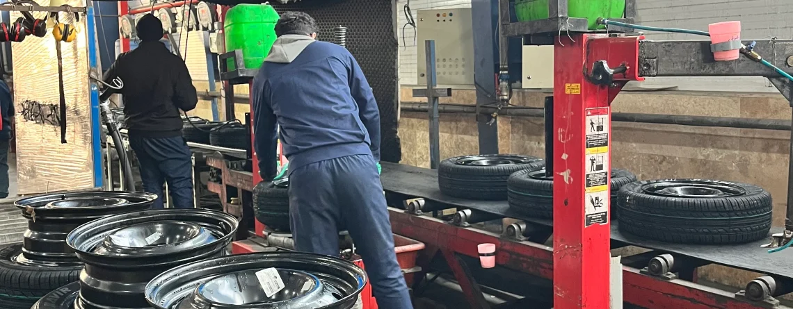 The image shows a tire assembly line in a factory setting. There are several tires mounted on rims placed on a conveyor belt. Two workers are seen in the background, wearing blue uniforms and working on the assembly process. The factory has high ceilings with blue-tinted windows, and various machinery and equipment are visible. The environment appears to be industrial, with a focus on tire manufacturing or assembly