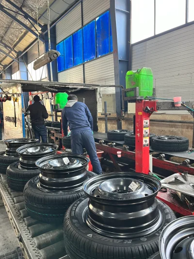 The image shows a tire assembly line in a factory setting. There are several tires mounted on rims placed on a conveyor belt. Two workers are seen in the background, wearing blue uniforms and working on the assembly process. The factory has high ceilings with blue-tinted windows, and various machinery and equipment are visible. The environment appears to be industrial, with a focus on tire manufacturing or assembly