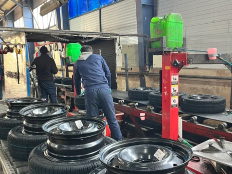 The image shows a tire assembly line in a factory setting. There are several tires mounted on rims placed on a conveyor belt. Two workers are seen in the background, wearing blue uniforms and working on the assembly process. The factory has high ceilings with blue-tinted windows, and various machinery and equipment are visible. The environment appears to be industrial, with a focus on tire manufacturing or assembly