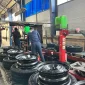 The image shows a tire assembly line in a factory setting. There are several tires mounted on rims placed on a conveyor belt. Two workers are seen in the background, wearing blue uniforms and working on the assembly process. The factory has high ceilings with blue-tinted windows, and various machinery and equipment are visible. The environment appears to be industrial, with a focus on tire manufacturing or assembly