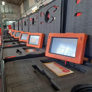 A row of diagnostic devices labeled 'MOTORSCAN' are meticulously arranged in protective foam cases. Each device has an orange casing with a screen, accompanied by red connectors and cables in the foam cutouts. The background suggests an industrial or workshop environment with high ceilings and fluorescent lighting