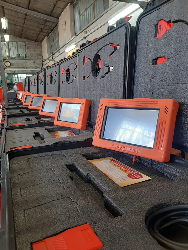 A row of diagnostic devices labeled 'MOTORSCAN' are meticulously arranged in protective foam cases. Each device has an orange casing with a screen, accompanied by red connectors and cables in the foam cutouts. The background suggests an industrial or workshop environment with high ceilings and fluorescent lighting