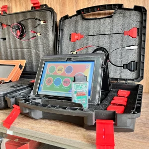 In a well-organized toolkit, an array of electronic diagnostic instruments eagerly awaits use. Nestled in a sleek black case, various compartments hold intricate devices and cables with red and black connectors. At the center, a vibrant tablet-like device showcases colorful icons—depicting a car, wrench, star, and gears—hinting at its multifaceted functions. This diagnostic ensemble, resting on a wooden surface, is surrounded by other intriguing items, suggesting a workshop brimming with potential discoveries and solutions