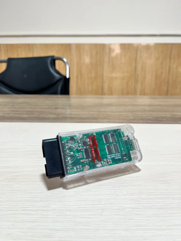 A close-up of an OBD-II scanner with a transparent plastic casing, revealing the internal circuit board and components. The text 'OBDII' and 'Smart' is visible on the circuit board. The device is plugged into a wooden surface, with a black chair and a wooden wall in the background