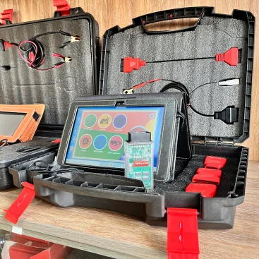 The image shows an open black plastic case with red latches containing various electronic diagnostic tools and cables. The case is divided into compartments with foam padding to securely hold the items. Inside the case, there are several cables with different connectors, including red and black clips, and a tablet-like device with a colorful interface displaying icons for different functions. The tablet is connected to a circuit board with a green PCB and various electronic components. The case also contains additional red connectors and a black zippered pouch. The setup appears to be a diagnostic tool kit, likely used for automotive or electronic diagnostics, making it relevant for technicians or engineers who need to troubleshoot and repair electronic systems