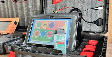 The image shows an open black plastic case with red latches containing various electronic diagnostic tools and cables. The case is divided into compartments with foam padding to securely hold the items. Inside the case, there are several cables with different connectors, including red and black clips, and a tablet-like device with a colorful interface displaying icons for different functions. The tablet is connected to a circuit board with a green PCB and various electronic components. The case also contains additional red connectors and a black zippered pouch. The setup appears to be a diagnostic tool kit, likely used for automotive or electronic diagnostics, making it relevant for technicians or engineers who need to troubleshoot and repair electronic systems