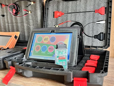 The image shows an open black plastic case with red latches containing various electronic diagnostic tools and cables. The case is divided into compartments with foam padding to securely hold the items. Inside the case, there are several cables with different connectors, including red and black clips, and a tablet-like device with a colorful interface displaying icons for different functions. The tablet is connected to a circuit board with a green PCB and various electronic components. The case also contains additional red connectors and a black zippered pouch. The setup appears to be a diagnostic tool kit, likely used for automotive or electronic diagnostics, making it relevant for technicians or engineers who need to troubleshoot and repair electronic systems