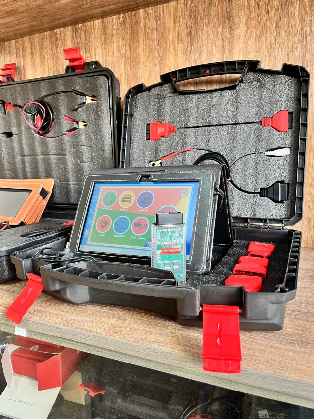 The image shows an open black plastic case with red latches containing various electronic diagnostic tools and cables. The case is divided into compartments with foam padding to securely hold the items. Inside the case, there are several cables with different connectors, including red and black clips, and a tablet-like device with a colorful interface displaying icons for different functions. The tablet is connected to a circuit board with a green PCB and various electronic components. The case also contains additional red connectors and a black zippered pouch. The setup appears to be a diagnostic tool kit, likely used for automotive or electronic diagnostics, making it relevant for technicians or engineers who need to troubleshoot and repair electronic systems