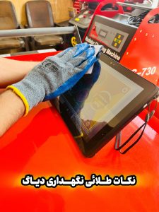 "A person wearing a blue and gray glove cleans a tablet screen with a cloth. The tablet rests on a red surface. In the background, there is a machine labeled 'Rebuilder Charging Machine' with the model number 'C-730.' Two chairs are also visible. The image includes Persian text that translates to 'Golden tips for maintaining diagnostics.