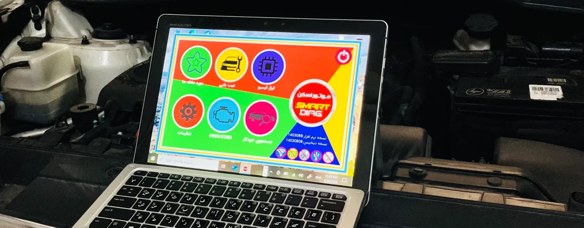 An open Kia vehicle hood with a laptop placed on the front grille. The laptop screen shows a colorful interface with various icons and Arabic text, likely related to vehicle diagnostics or maintenance