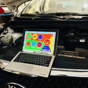 An open Kia vehicle hood with a laptop placed on the front grille. The laptop screen shows a colorful interface with various icons and Arabic text, likely related to vehicle diagnostics or maintenance