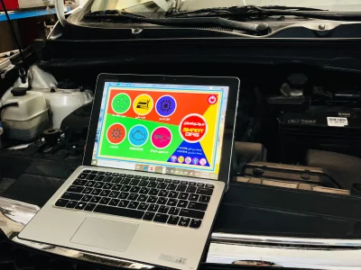 An open Kia vehicle hood with a laptop placed on the front grille. The laptop screen shows a colorful interface with various icons and Arabic text, likely related to vehicle diagnostics or maintenance