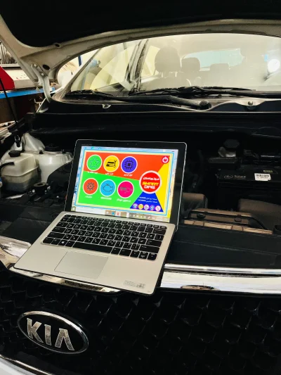 An open Kia vehicle hood with a laptop placed on the front grille. The laptop screen shows a colorful interface with various icons and Arabic text, likely related to vehicle diagnostics or maintenance